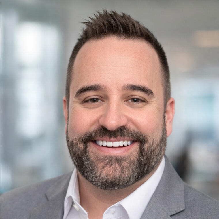 Micah of Micah Makes Cents headshot photo of Micah, from Micah Makes cents. Blurred background with his facial features in focus - smiling with a white collard shirt on under a taupe grey colored jacket.